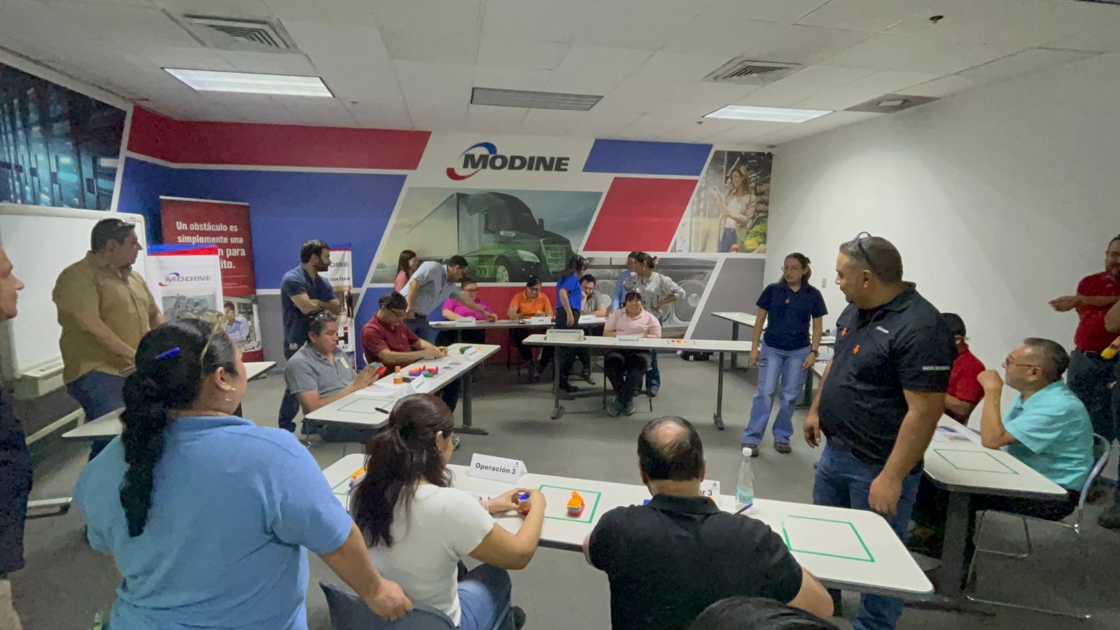 Modine employees take part in a hands-on team training exercise at tables labeled "Operación 3," working with colored pieces in a branded conference room with the Modine logo and Spanish-language signage on the walls.