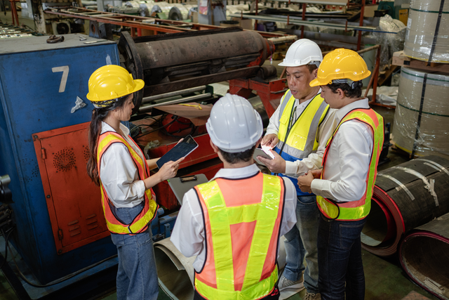 Four workers in hard hats and safety vests gather around a tablet, discussing something together on a factory floor beside industrial machinery.