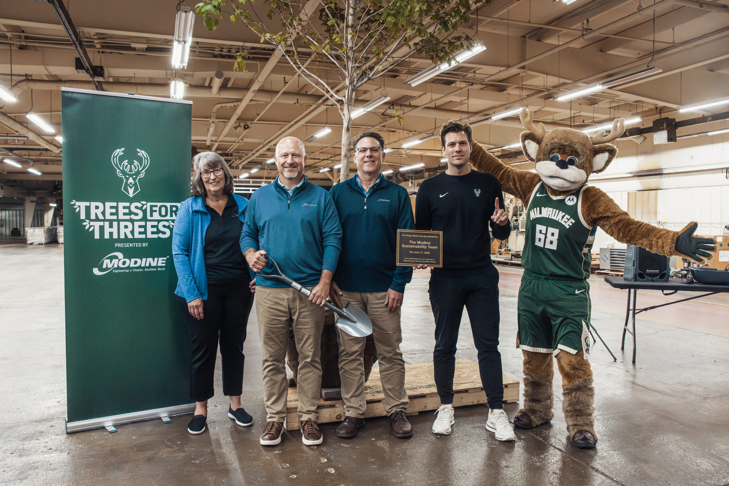 Modine team members pose with a newly planted tree, a shovel, and a commemorative plaque alongside the Milwaukee Bucks mascot at a "Trees for Threes" sustainability event, with a banner noting the program is presented by Modine.