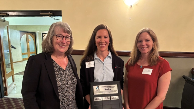 Three women smile while holding a Modine award plaque recognizing the company as a distinguished partner in education.