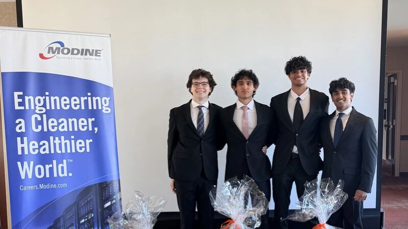 Four young men in dark suits stand together next to a Modine "Engineering a Cleaner, Healthier World" banner at a career event.