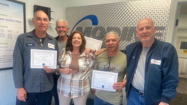 Five Modine employees smile together in an office, two holding certificates, beneath the company logo and mission statement on the wall.