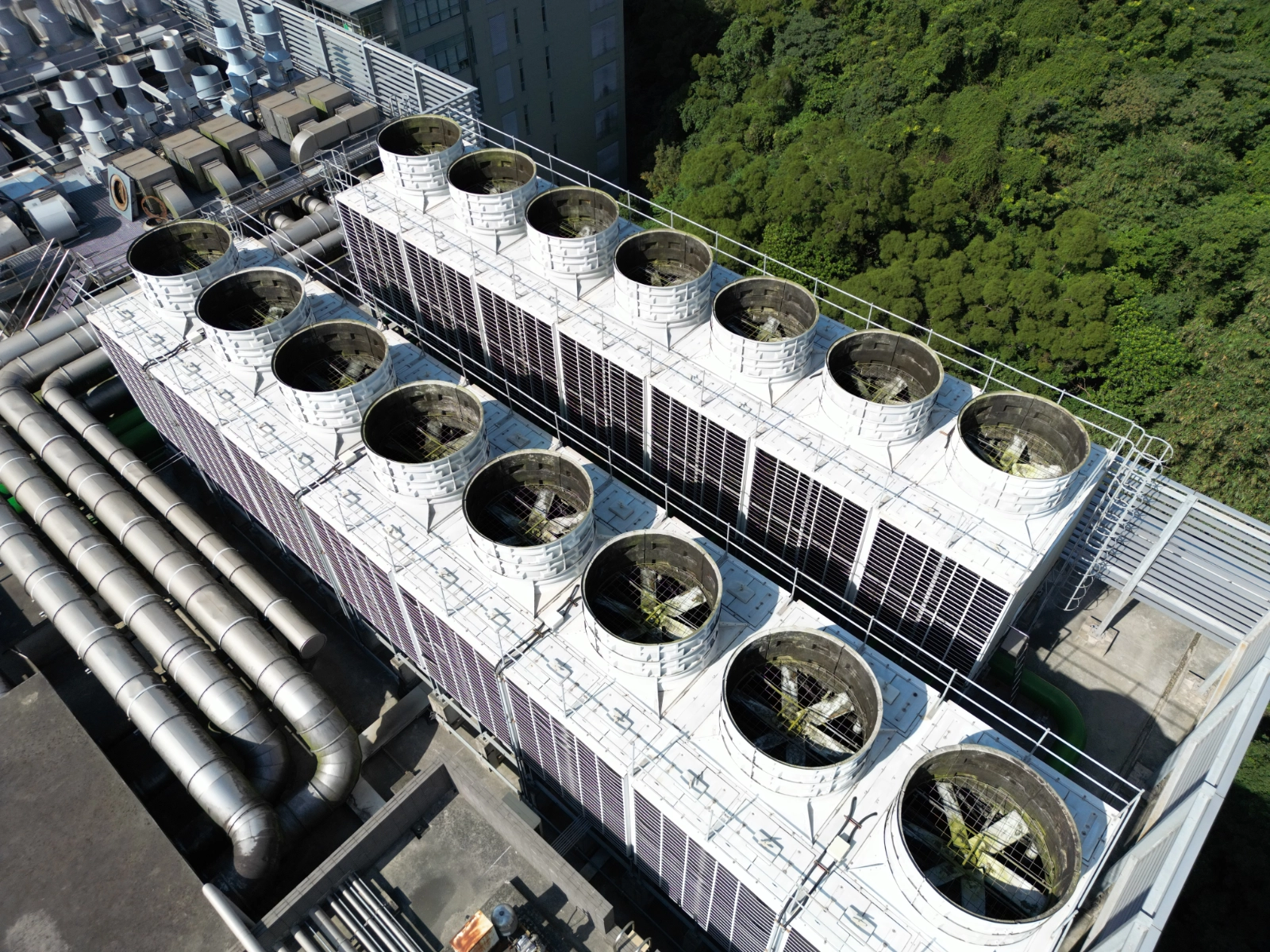 An aerial view of large industrial cooling towers with rows of circular fans on a building rooftop, with insulated pipes running alongside and green forest in the background.