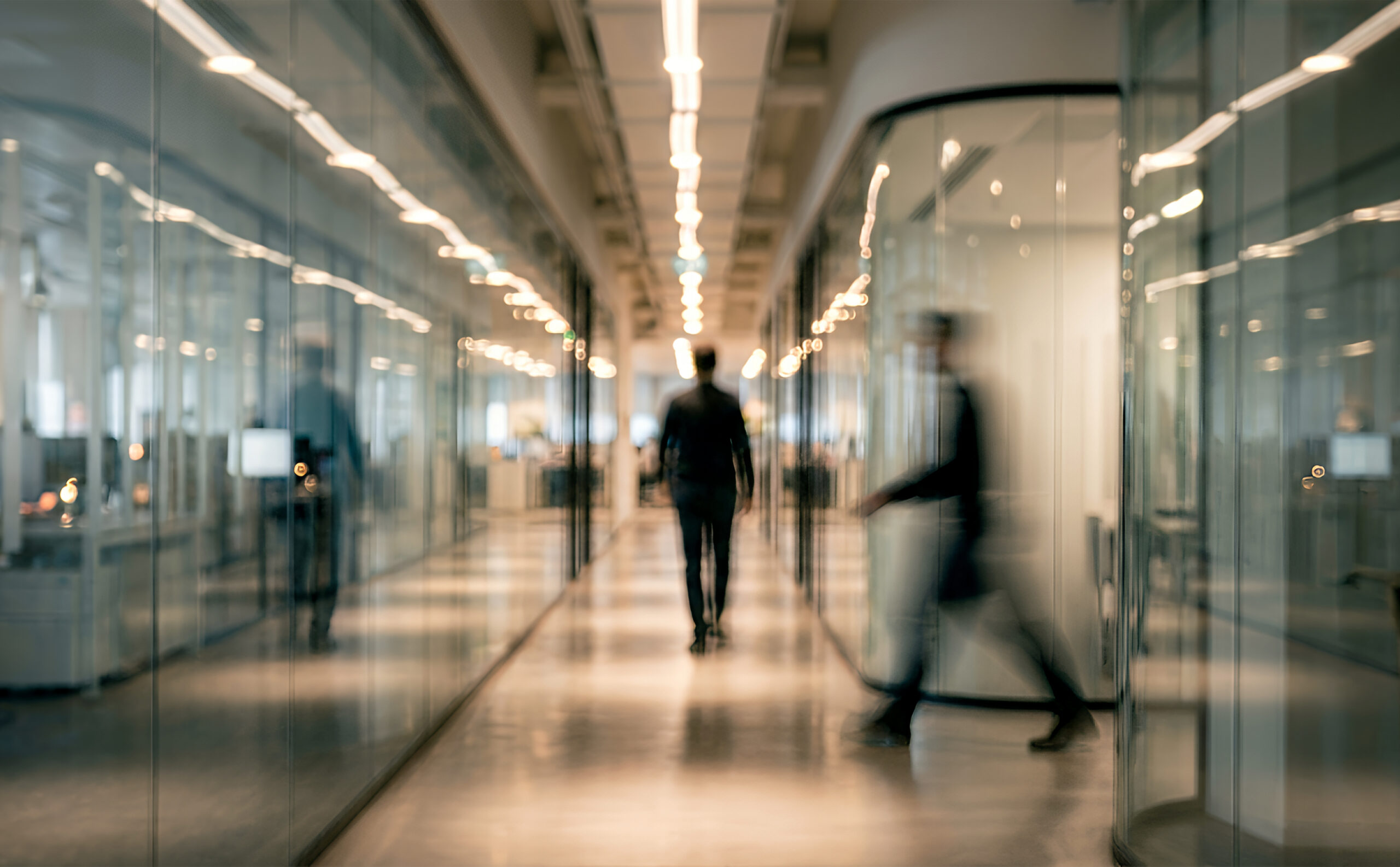 Blurred office hallway with glass-walled rooms and warm overhead lights, a person walking away and another passing in motion.