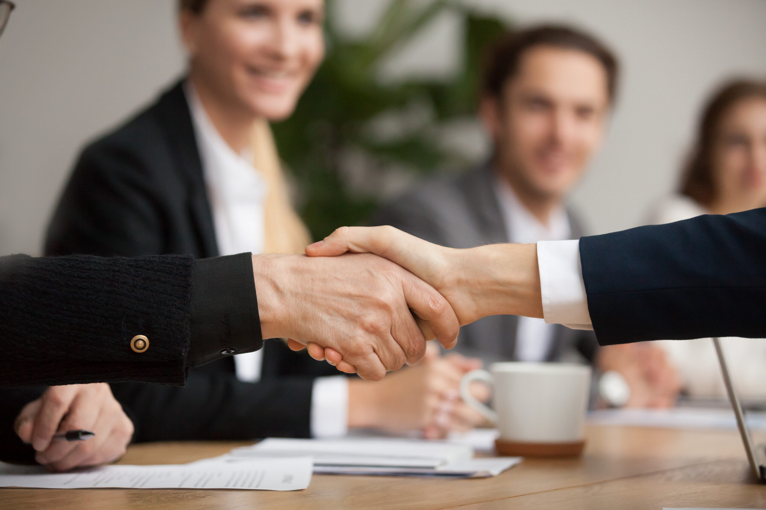 Two business professionals shake hands across a meeting table while smiling colleagues look on in the background.